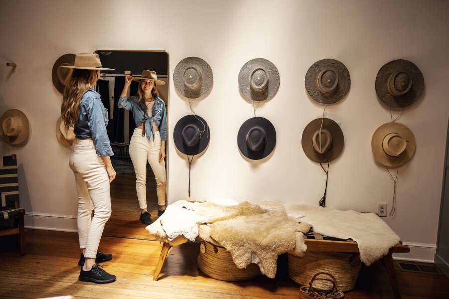 Woman looking into full-length mirror with hand on hat at a store with hats on the wall. 