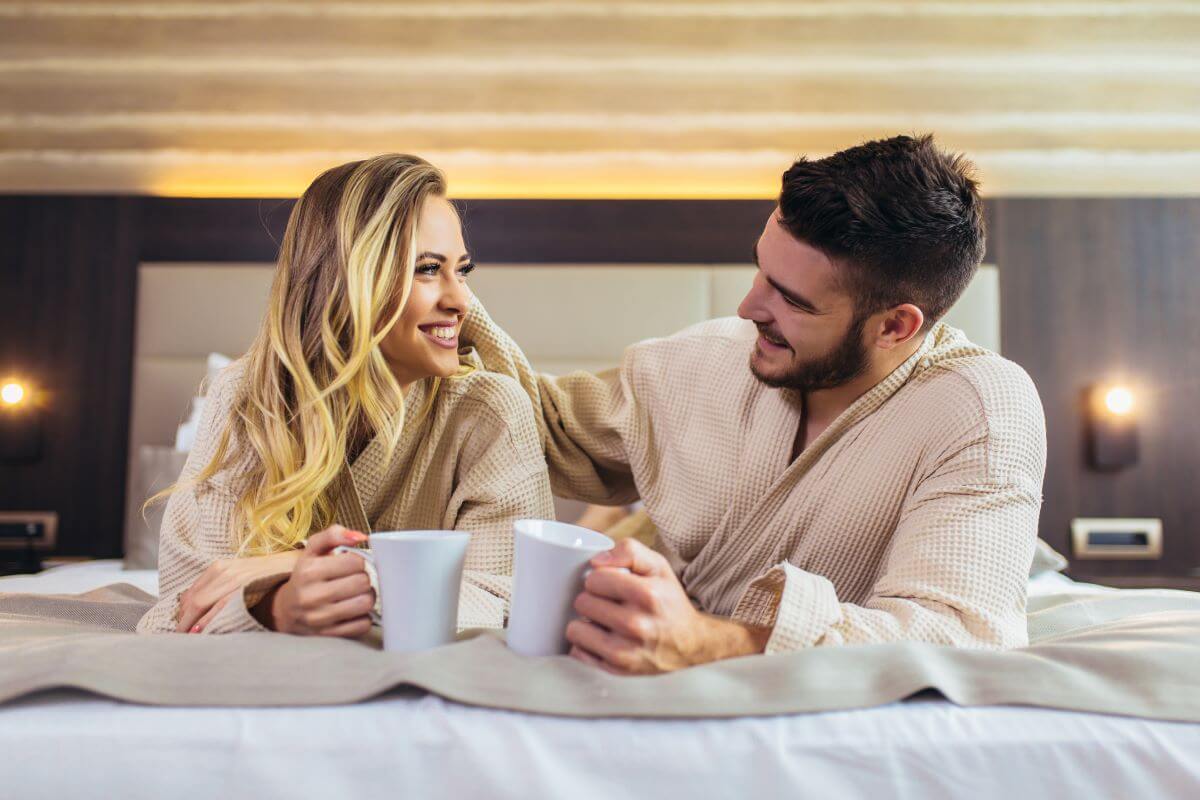 Couple propped on elbows on hotel bed facing smiling at each other, mugs in hands.