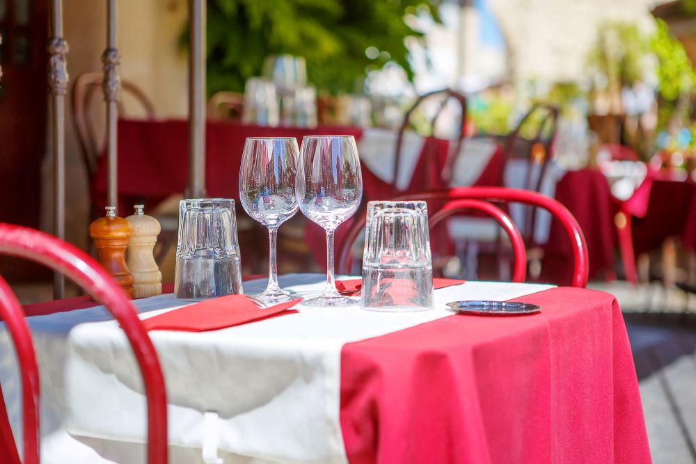 Sidewalk cafe table for 2 with red table cloth and white runner, napkins and wine and water glasses.