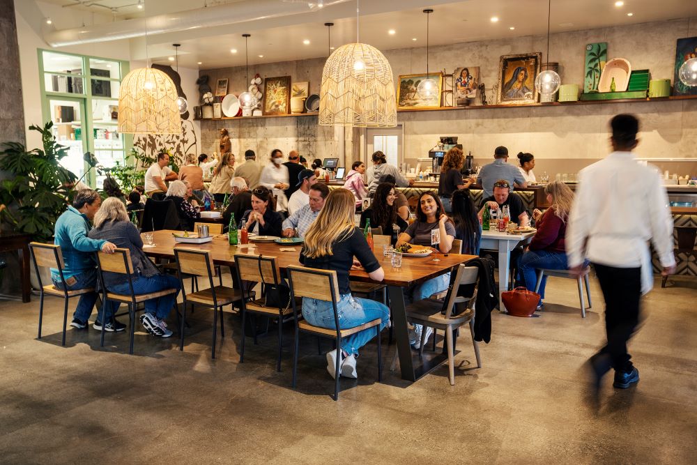 Diners at community tables under large chandeliers. Bar seating behind. Art on wall. 