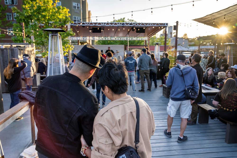 Man wearing fedora with short haired woman standing on deck with diners, watching band. Several tall outdoor heaters.  