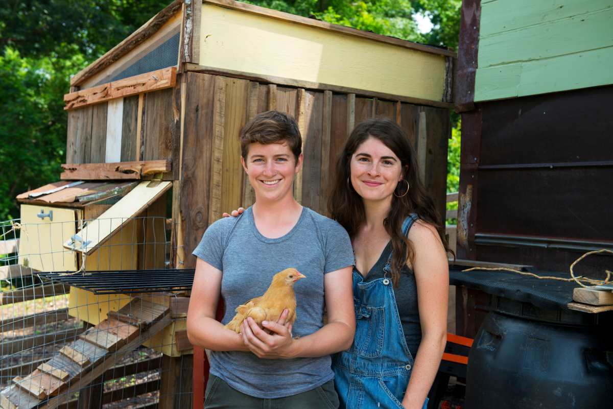 Two smiling women in front of chicken coup. Long-haired brunette in overalls with hand on should of woman holding hen.